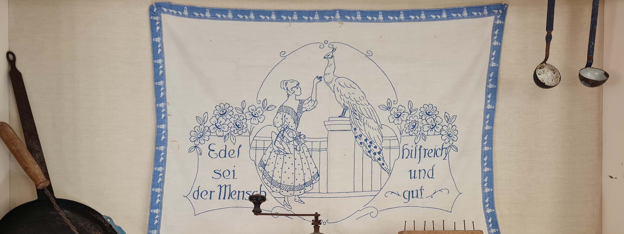 Hímzett terítő, mely egy nőt és egy pávát ábrázol mellette német felirattal